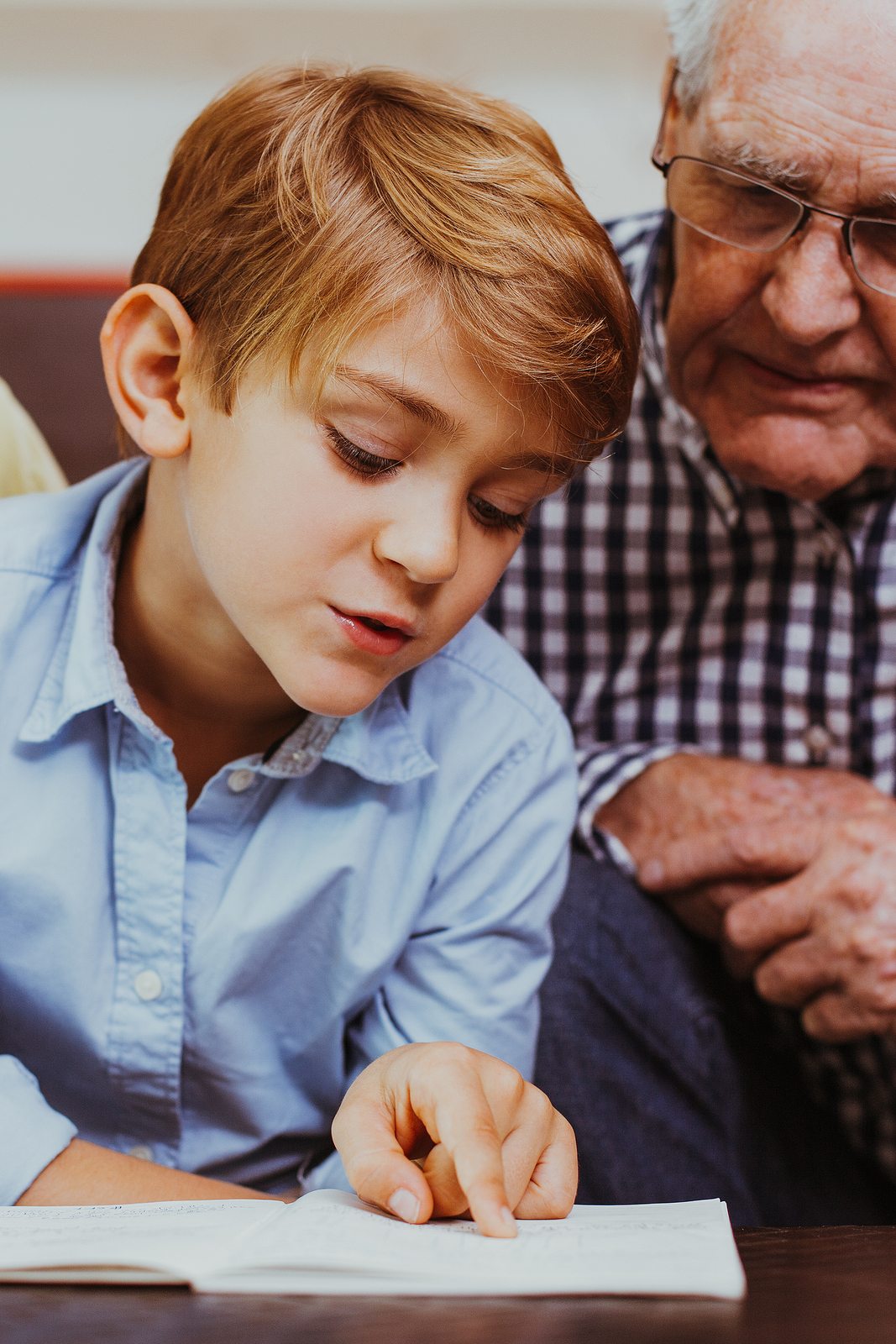 bigstock--216972124 Portrait of diligent little schoolboy sitting at table and reading book with grandparents touching pages. Senior man and woman wearing glasses looking at grandson and smiling